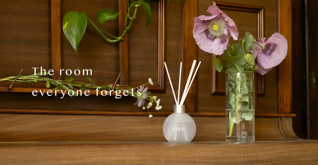 AIRY® Fragrances reed diffuser on a wooden console beside fresh pink poppies and trailing green leaves, styled as a considered home entrance,  luxury home fragrance by AIRY® Fragrances.