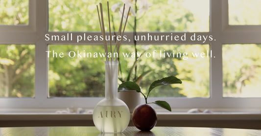 AIRY® Fragrances reed diffuser on a low wooden surface beside a fresh peach and botanical leaf, with warm natural light and green trees through an open window, evoking the Okinawan philosophy of slow living and daily ritual.