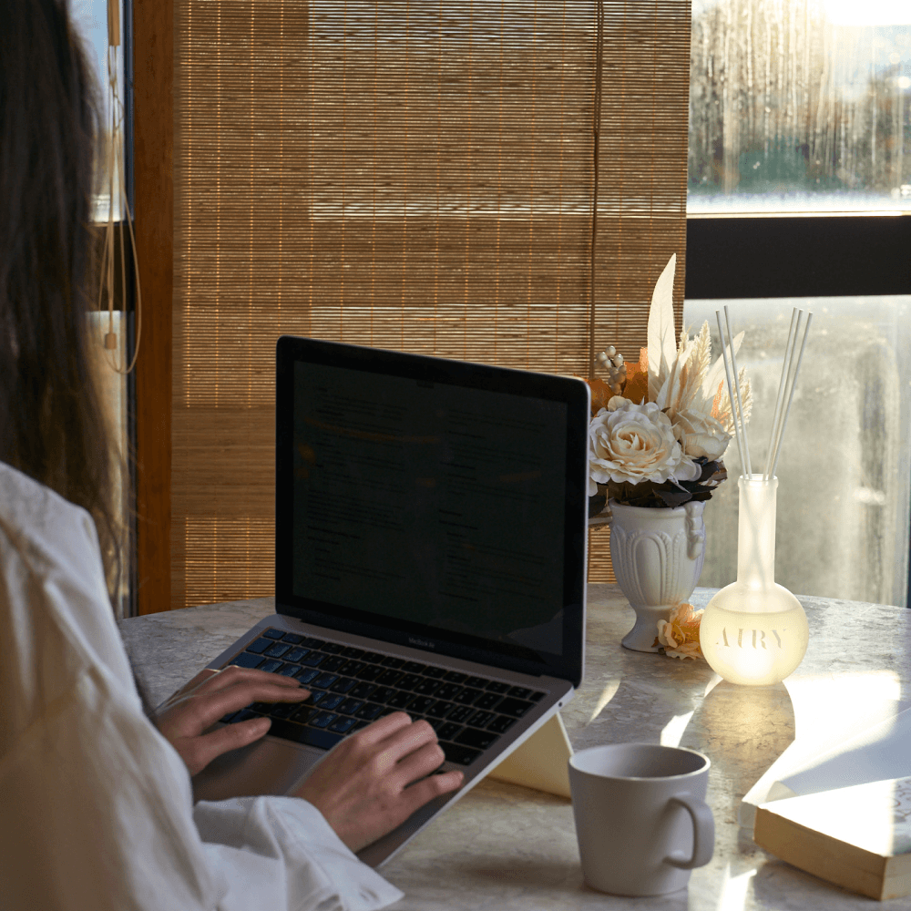 A woman working at a laptop in a sunlit home office with the Serene Solstice 寧 reed diffuser on the desk. The image highlights the grounding Chiba Rose and Frankincense aroma, designed to create a calm and focused atmosphere for remote work. An ideal Japanese luxury fragrance for modern desks and creative studios.