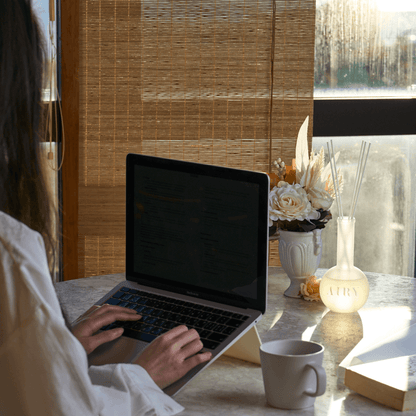 A woman working at a laptop in a sunlit home office with the Serene Solstice 寧 reed diffuser on the desk. The image highlights the grounding Chiba Rose and Frankincense aroma, designed to create a calm and focused atmosphere for remote work. An ideal Japanese luxury fragrance for modern desks and creative studios.