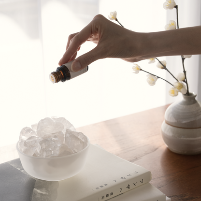 Demonstration of the Airy Fragrances scent ritual: dripping Blissful Harmony 悦 (Etsu) fragrant oil onto raw Clear Quartz stones. Styled in a minimalist home setting on a wooden table with design books and a ceramic vase. This heat-free Japanese method uses alcohol-free botanical oil to create a fresh, localized atmosphere of joy and mental clarity.