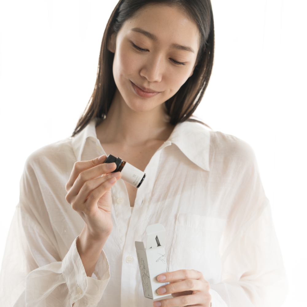 A woman happily unboxing the Airy Fragrances Blissful Harmony 悦 (Etsu) fragrant oil. She holds the 10ml amber bottle next to its open white box, showcasing a moment of quiet wellness and joyful anticipation. This Japanese-crafted, alcohol-free blend of green tea and gardenia is designed to elevate the home atmosphere, especially when paired with the Airy Clear Quartz Stone Diffuser
