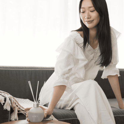 Serene lifestyle portrait of a woman resting on a sofa with the Elegant Harmony 優 reed diffuser. The image captures the frosted glass vessel and white reeds in a minimalist setting, evoking the poised, quiet luxury and balanced atmosphere of Kyoto Osmanthus and White Tea.