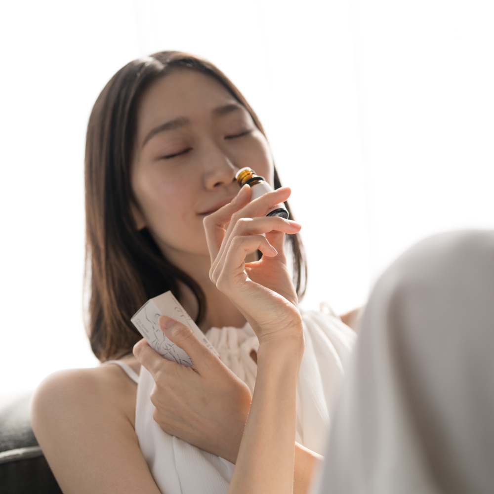 Lifestyle image of a woman happily inhaling the scent from an Airy Fragrances Elegant Harmony 優 (Yu) oil bottle. She holds the 10ml amber bottle and its minimalist box, showcasing a moment of quiet wellness, composed serenity, and mental clarity. This Japanese-crafted, alcohol-free blend of white tea and golden osmanthus is designed to ground the home atmosphere, especially when paired with the Airy Clear Quartz Stone Diffuser