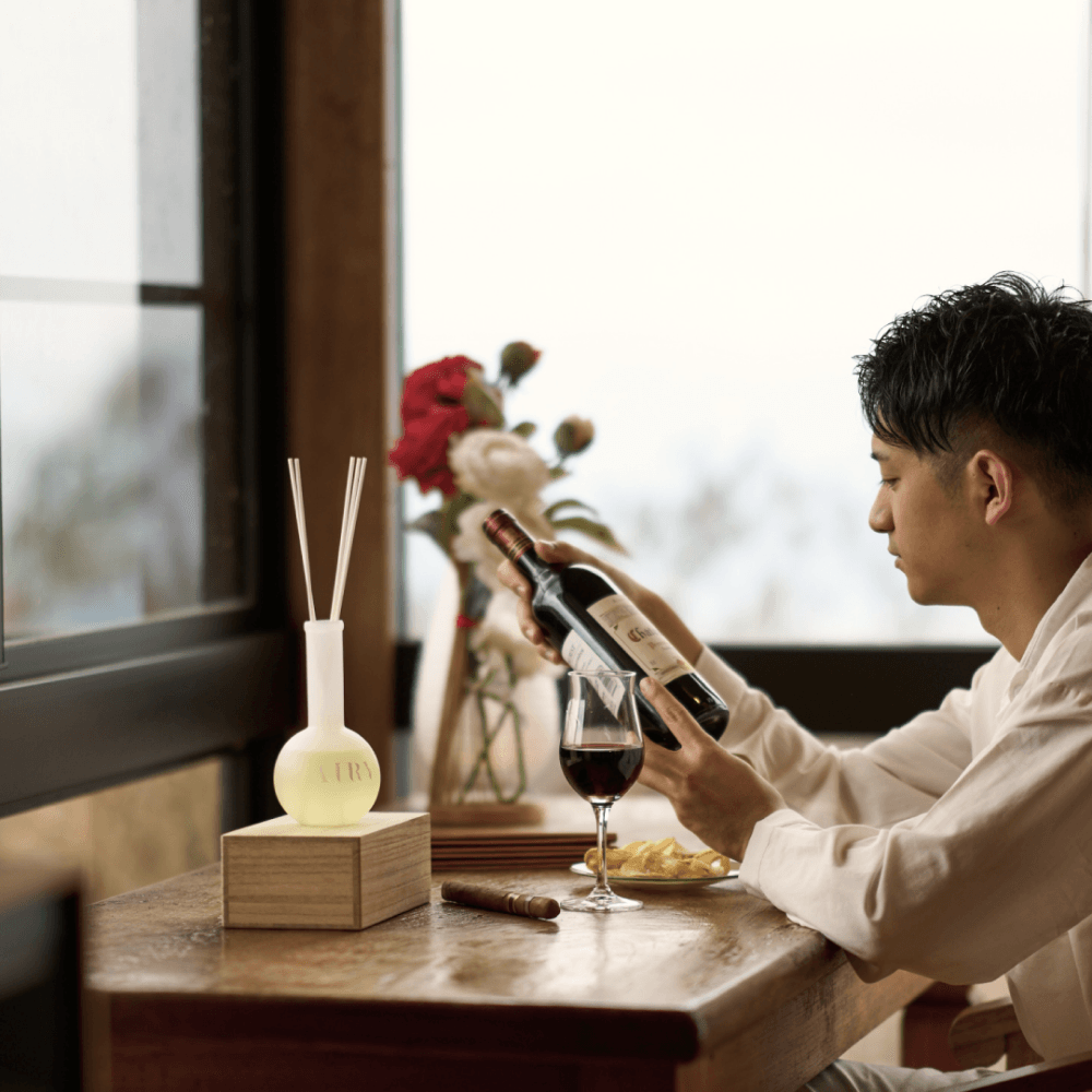 Masculine luxury lifestyle setting for Forest Breath 森 reed diffuser. A man in a refined interior enjoying a glass of wine, with the Nagano Hinoki diffuser prominently displayed. This visual reinforces the woody, grounding character of the scent, making it a perfect gift for men and sophisticated dens or home offices