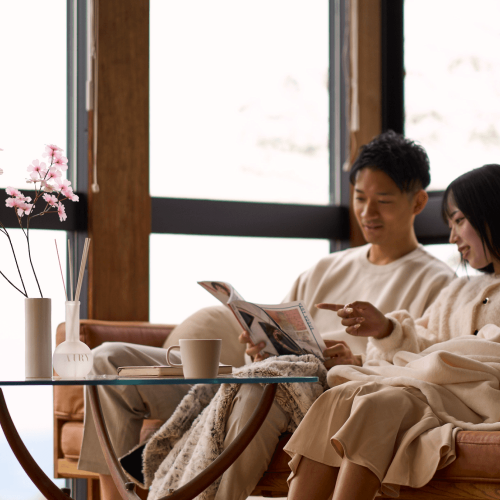 A couple in a refined, minimalist living room enjoying the uplifting aroma of Mystic Essence 空. The frosted glass reed diffuser and cherry blossom branches are styled on a glass coffee table, highlighting the fresh, dewy Tokyo Sakura scent. An ideal home fragrance for creating a bright, shared atmosphere in premium UK homes.