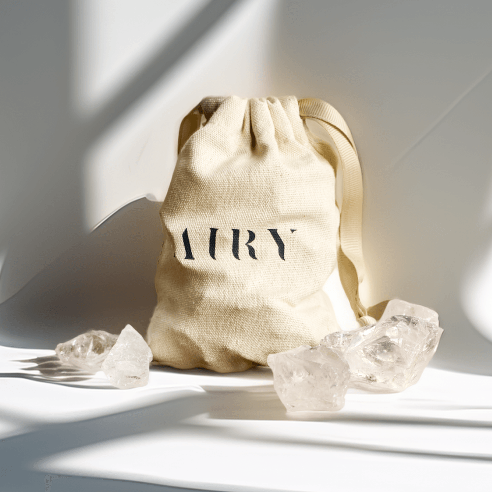 A natural, undyed cotton drawstring bag with the Airy Fragrances logo, from the Clear Quartz Stone Diffuser set. The multifunctional, unisex bag is shown next to the raw quartz stones, highlighting the premium, sustainable, and reusable Japanese-crafted packaging included in the Editor's Pick wellness set.
