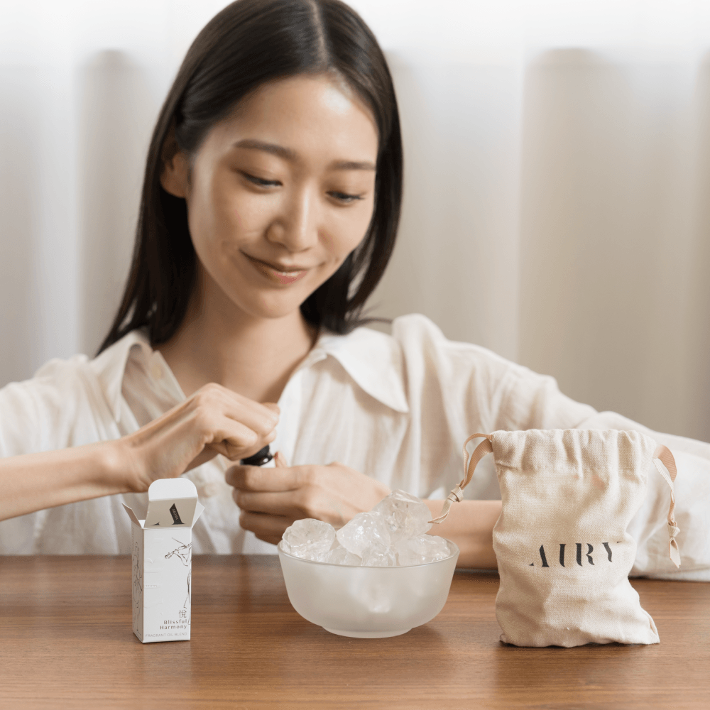 A woman performing the Airy Fragrances scent ritual, opening a 10ml bottle of Blissful Harmony 悅 (Etsu) oil to apply to the Clear Quartz Stone Diffuser. Set on a wooden table with the signature cotton bag, this image demonstrates the simple, heat-free way to refresh home atmospheres with alcohol-free Japanese fragrance oils