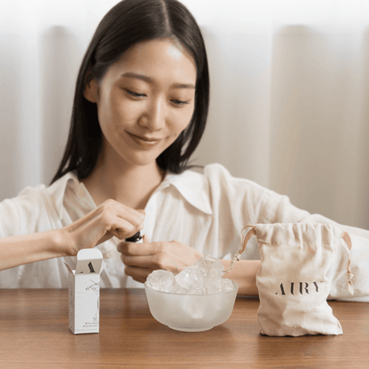 A woman performing the Airy Fragrances scent ritual, opening a 10ml bottle of Blissful Harmony 悅 (Etsu) oil to apply to the Clear Quartz Stone Diffuser. Set on a wooden table with the signature cotton bag, this image demonstrates the simple, heat-free way to refresh home atmospheres with alcohol-free Japanese fragrance oils