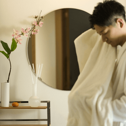 Morning wellness ritual featuring Sweet Infusion 幻 luxury reed diffuser. A man in a bathrobe in a minimalist bathroom setting, with the frosted glass diffuser and Magnolia blossoms displayed on a vanity shelf. Captures the fresh, tea-inspired serenity ideal for bathrooms and start-of-day relaxation.