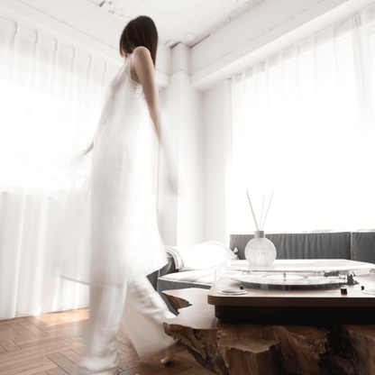 Elegant lifestyle scene in a sun-lit minimalist room featuring the Blissful Harmony 悦 reed diffuser on a rustic wooden table next to a record player. A woman in airy white clothing moves through the serene space, capturing the brand's calm and sophisticated Japanese aesthetic.