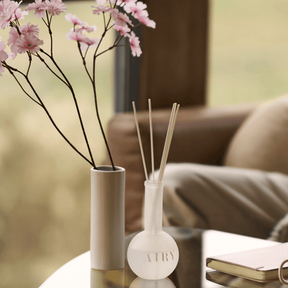 Mystic Essence 空 reed diffuser in a sunlit living room or reading nook. The frosted glass vessel is styled on a coffee table next to a branch of cherry blossoms and a notebook, highlighting the uplifting Tokyo Sakura scent. Perfect for creating a calm, creative atmosphere in modern home interiors.
