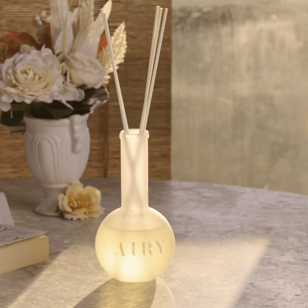 Serene Solstice 寧 luxury reed diffuser styled on a stone table during golden hour. The frosted glass bottle is surrounded by soft sunlight and white roses, highlighting the grounding Chiba Rose and Frankincense aroma. Perfect for creating a calm, atmospheric evening in modern UK living rooms and conservatories.