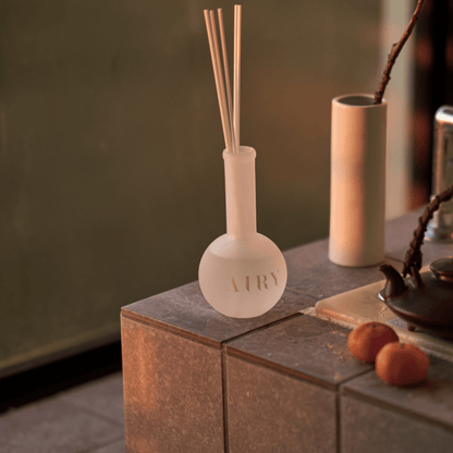Sweet Infusion 幻 reed diffuser in a serene Japanese spa setting. The frosted glass vessel is placed on stone tiling next to a traditional iron tea kettle, capturing the calming essence of Hakone Jin Xuan tea. A luxury home fragrance choice for bathrooms and wellness spaces.