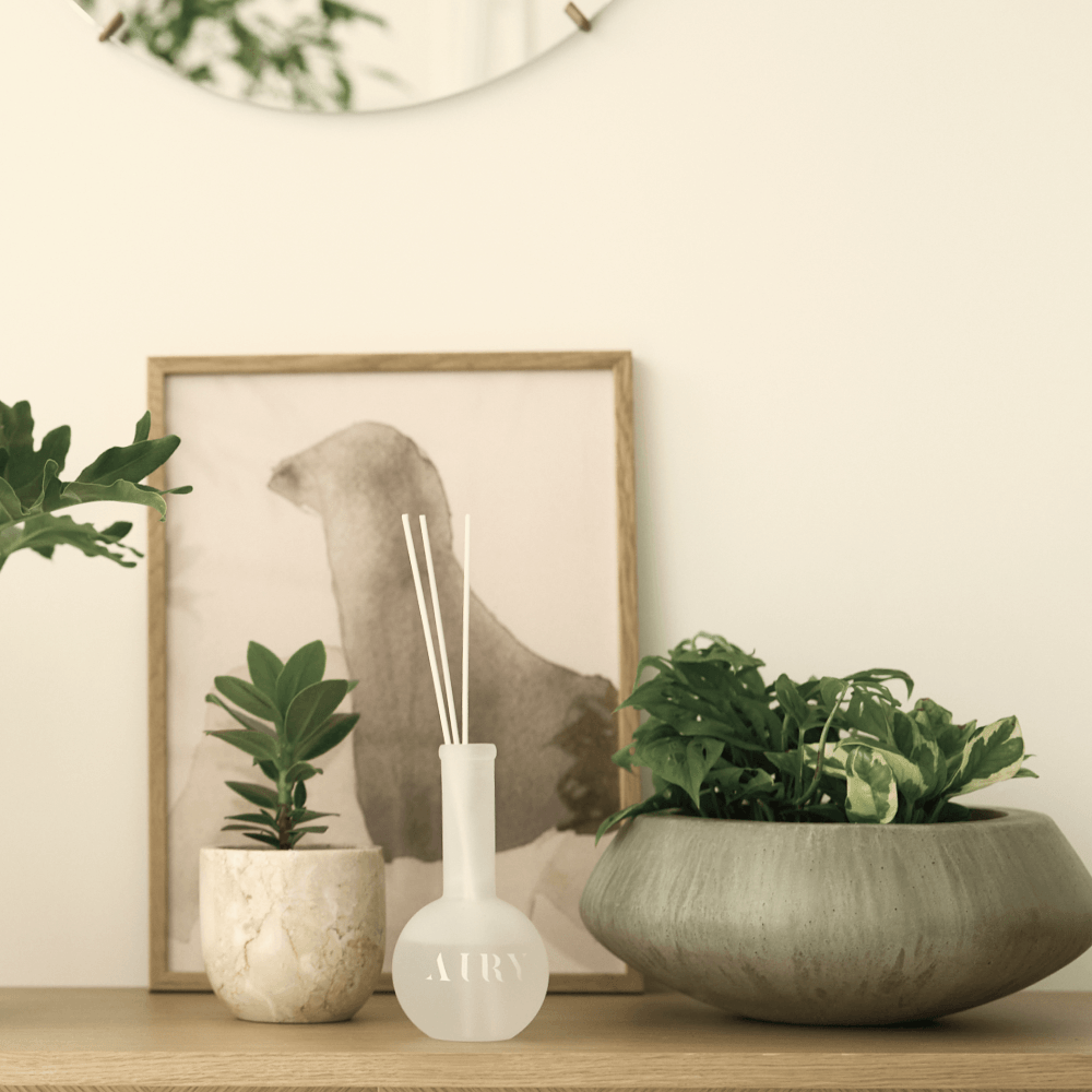 Velvet Embrace 韻 luxury reed diffuser styled in a minimalist Japanese home interior. The frosted glass vessel is placed on a wooden console next to lush green plants and abstract art, highlighting the silken Okayama Peach and Jasmine scent. Ideal for creating a calm, poetic atmosphere in modern living rooms and studios.