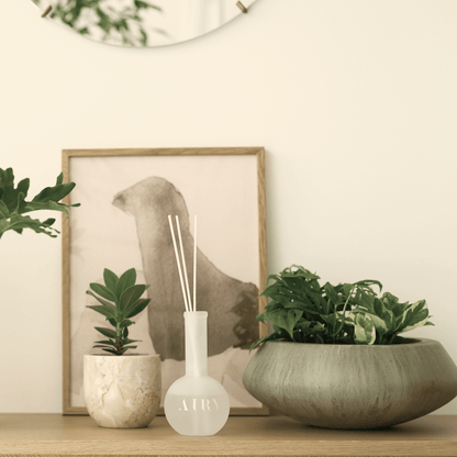 Velvet Embrace 韻 luxury reed diffuser styled in a minimalist Japanese home interior. The frosted glass vessel is placed on a wooden console next to lush green plants and abstract art, highlighting the silken Okayama Peach and Jasmine scent. Ideal for creating a calm, poetic atmosphere in modern living rooms and studios.